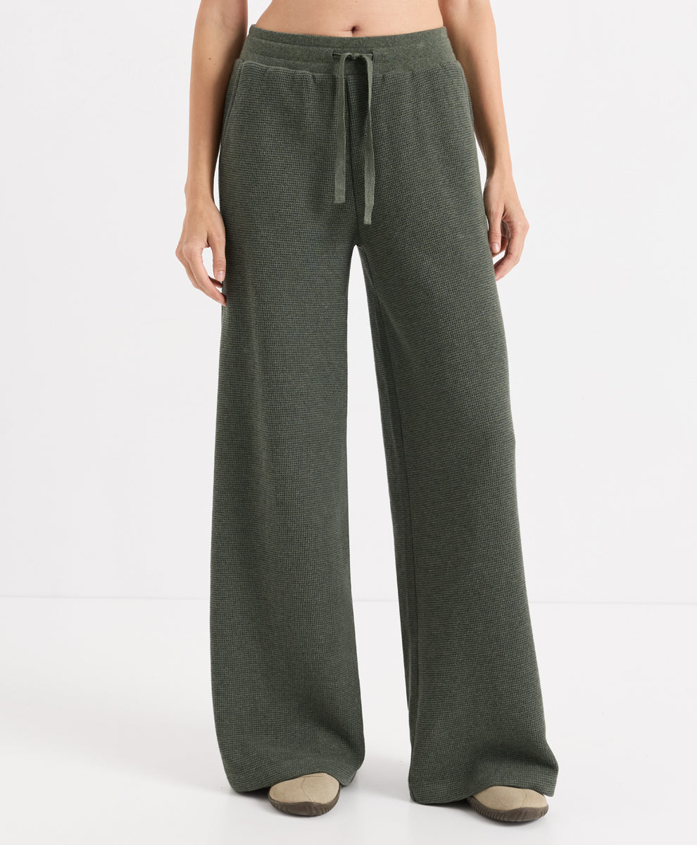 A model wearing a pair of wide-leg, dusted green heather pants with an elastic waistband and drawstring, paired with beige slip-on shoes against a plain white studio background.