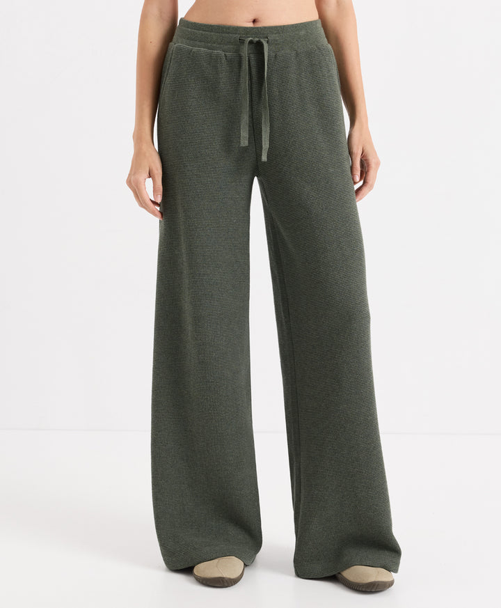 A model wearing a pair of wide-leg, dusted green heather pants with an elastic waistband and drawstring, paired with beige slip-on shoes against a plain white studio background.