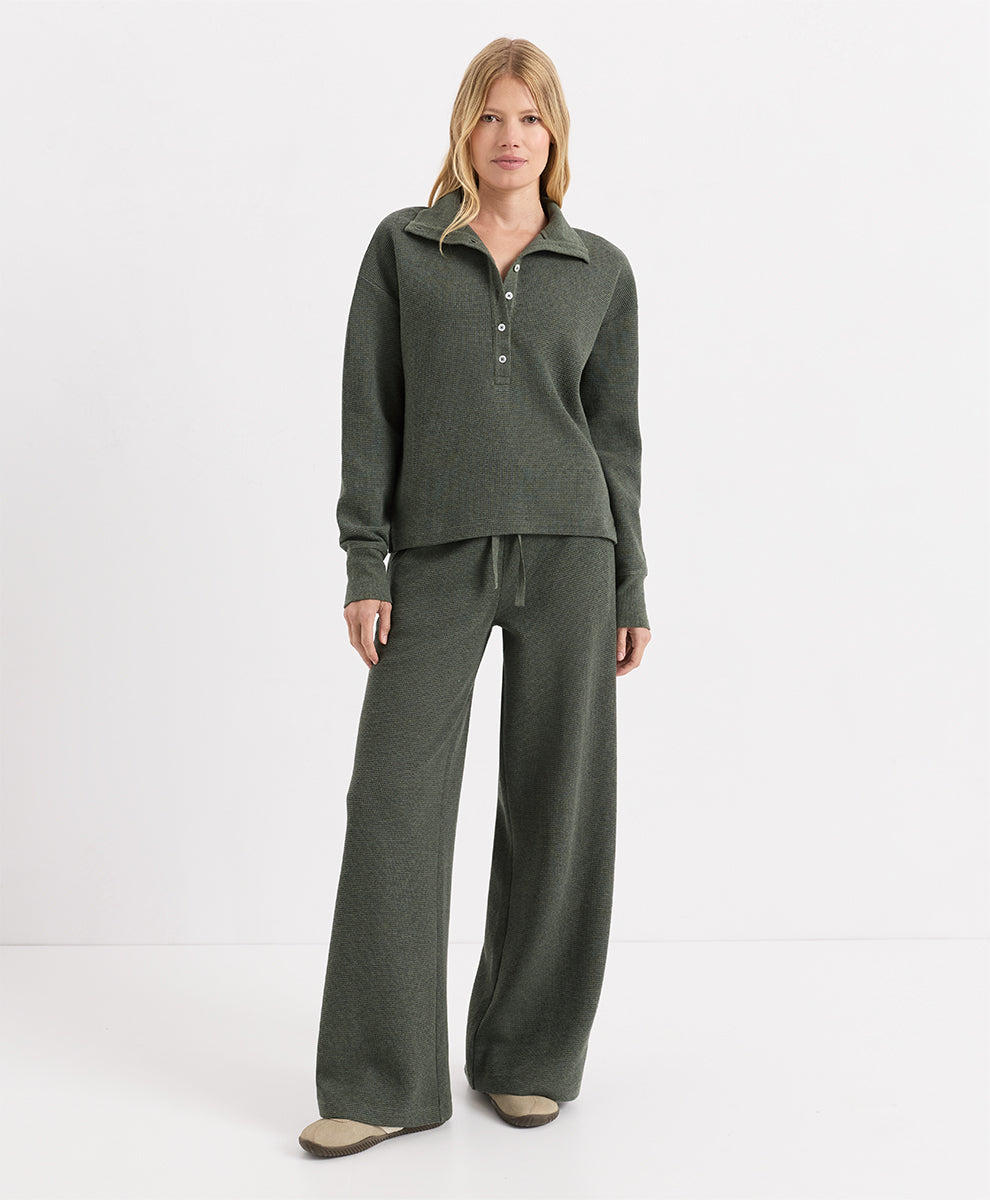 A woman modeling a matching two-piece knit loungewear set in dusted green heather, featuring a long-sleeve henley top with a button placket and matching wide-leg pants, against a plain white studio background.