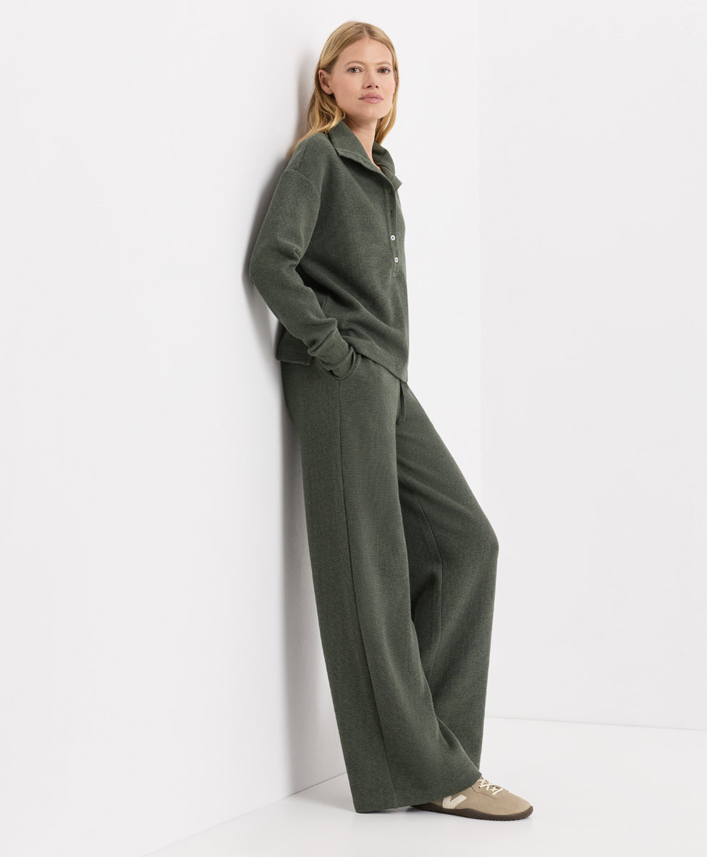 A woman stands against a white wall wearing a dusted green heather knit matching set consisting of a collared henley top and wide-leg pants, paired with beige loafers.