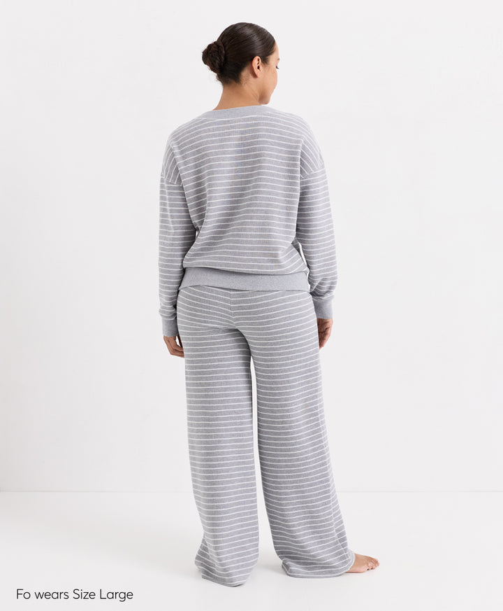 A woman stands with her back to the camera wearing a light gray and white striped long-sleeve top and matching wide-leg pants. The text &