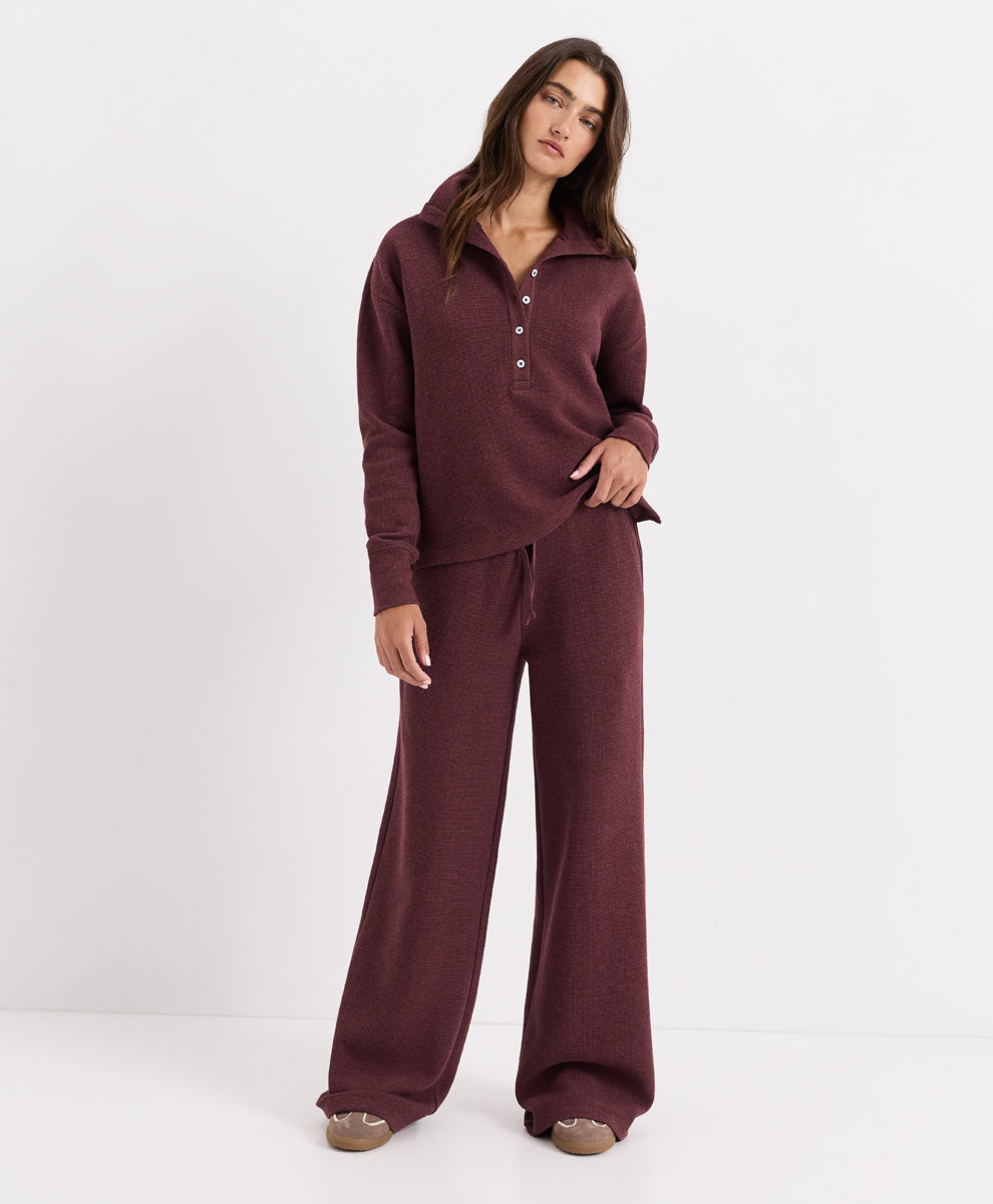 A woman modeling a matching merlot heather lounge set consisting of a long-sleeve henley top with a button placket and wide-leg pants, photographed against a clean white studio background.