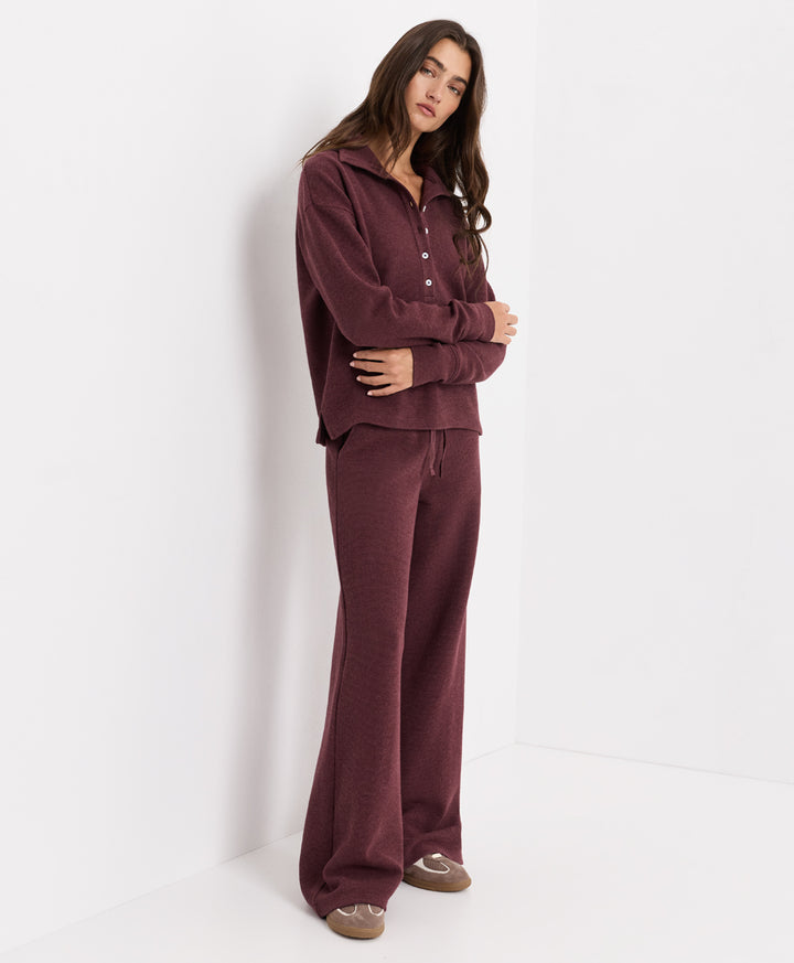 A woman modeling a matching merlot heather lounge set consisting of a long-sleeve button-front top and wide-leg pants, standing against a clean white studio background.