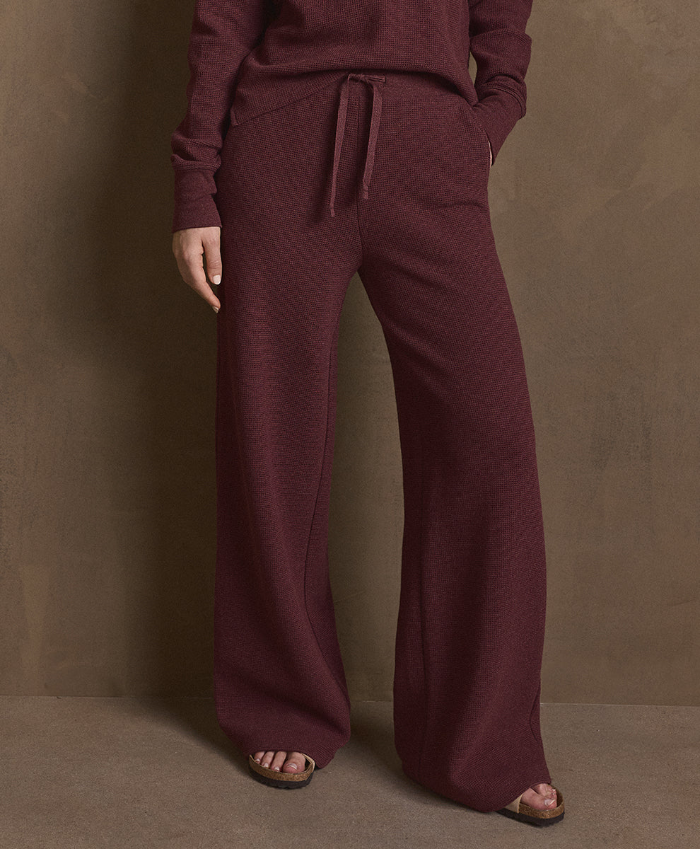 A person wearing a matching merlot heather knit lounge set with wide-leg drawstring pants and a long-sleeve top, standing against a textured brown studio backdrop.