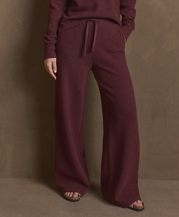 A person wearing a matching merlot heather knit lounge set with wide-leg drawstring pants and a long-sleeve top, standing against a textured brown studio backdrop.