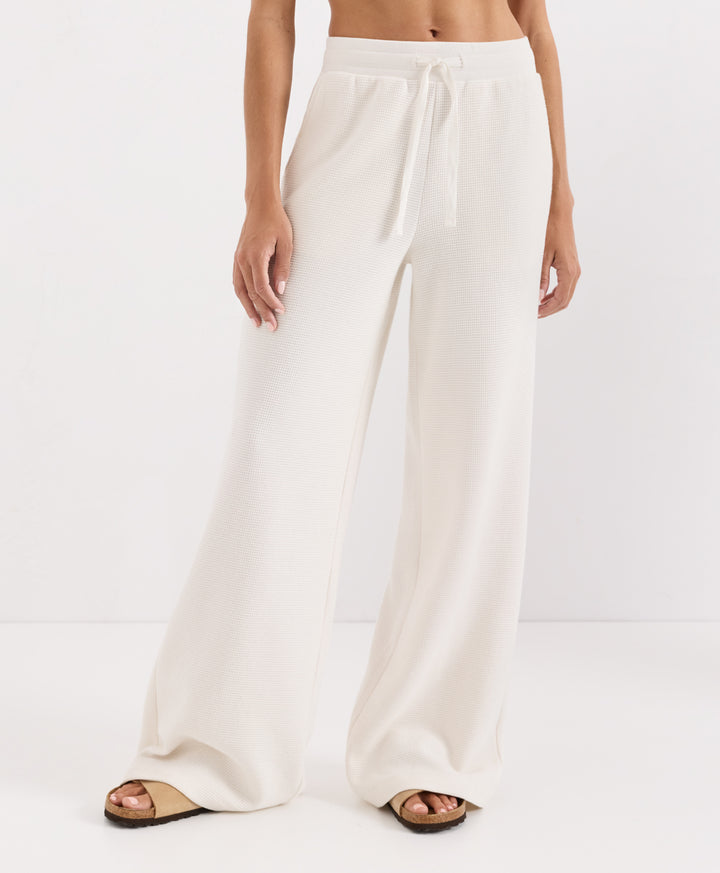 A model wearing high-waisted, wide-leg soft ivory pants with a drawstring waistband, paired with tan slide sandals, shown from the waist down against a clean white studio background.