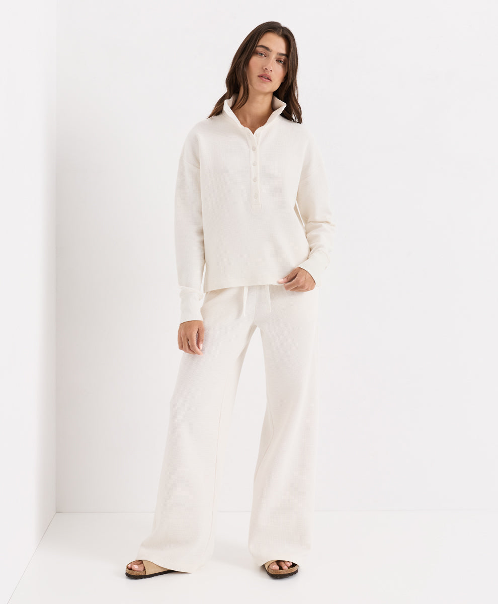 A woman modeling a matching soft ivory loungewear set consisting of a long-sleeve buttoned henley shirt and wide-leg pants, paired with dark brown slide sandals, set against a clean white studio background.