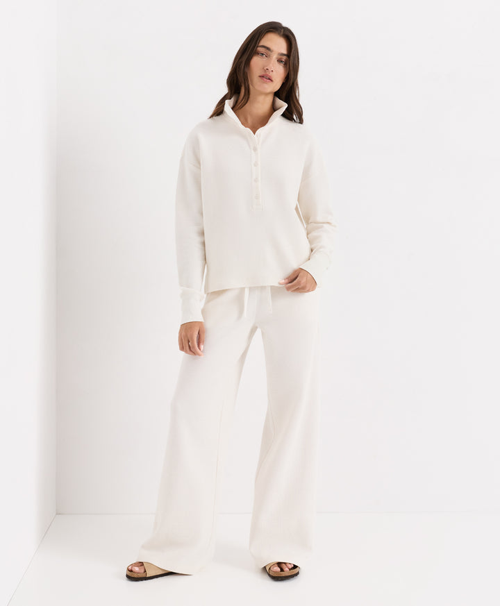 A woman modeling a matching soft ivory loungewear set consisting of a long-sleeve buttoned henley shirt and wide-leg pants, paired with dark brown slide sandals, set against a clean white studio background.