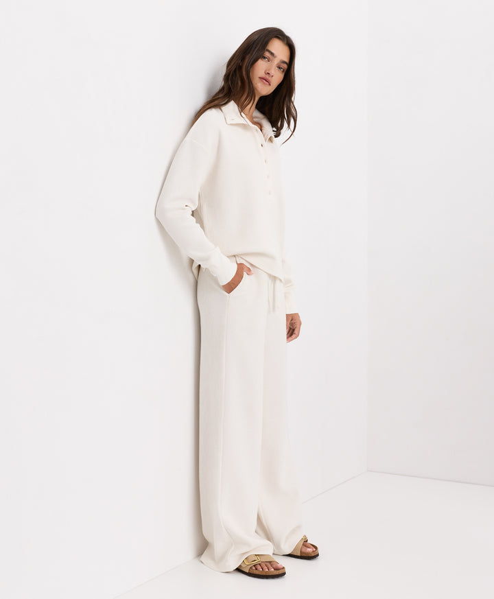 A woman stands in a minimalist white studio corner wearing a matching soft ivory lounge set consisting of a long-sleeve henley top and wide-leg pants, paired with brown sandals.