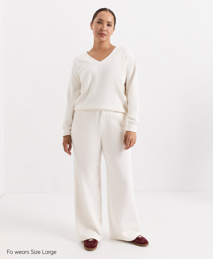 A woman modeling a matching soft ivory knit loungewear set consisting of a long-sleeve V-neck top and wide-leg pants, paired with burgundy slip-on shoes. The text &