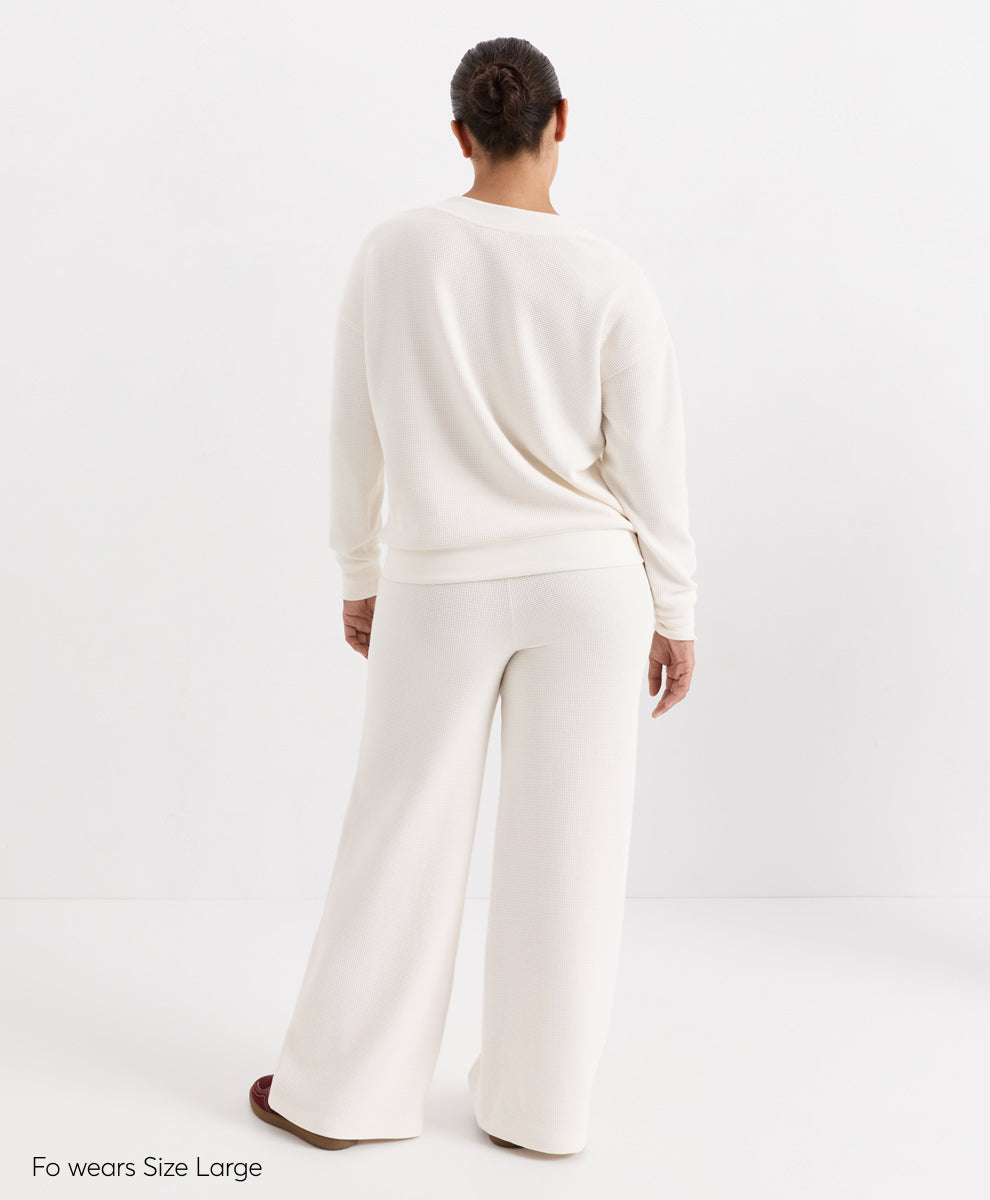 A model viewed from behind wearing a soft ivory V-neck top and matching wide-leg pants, standing against a plain white studio background. Text in the bottom-left corner reads &