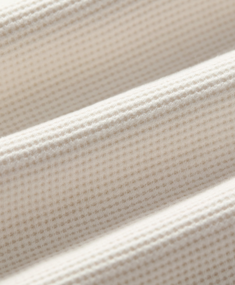 Close-up of soft ivory textured fabric with a fine waffle or grid-like weave, shown in soft diagonal folds.