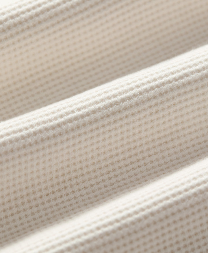 Close-up of soft ivory textured fabric with a fine waffle or grid-like weave, shown in soft diagonal folds.