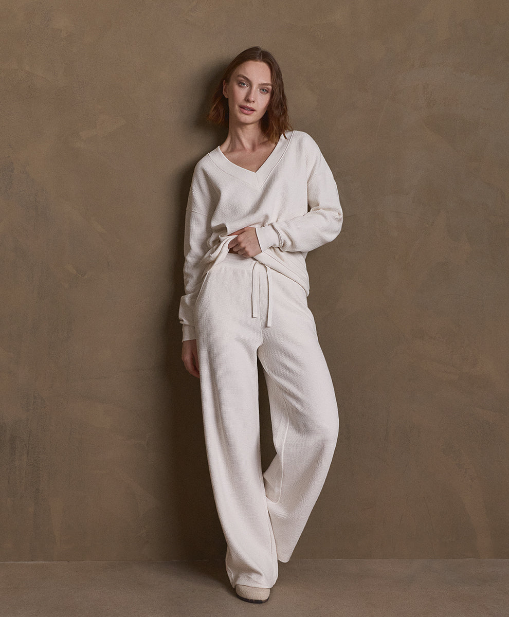 A woman models a matching soft ivory lounge set consisting of a long-sleeve V-neck top and wide-leg pants, standing against a textured brown studio backdrop.