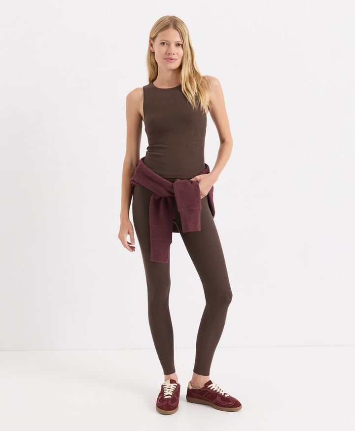 A woman modeling a sleeveless cocoa ribbed tank top, matching leggings, and burgundy sneakers, with a burgundy knit sweater tied around her waist, against a plain white studio background.