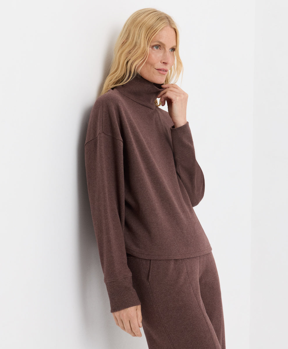 A blonde woman wearing a matching chestnut heather set with a funnel neck long-sleeve top and pants, leaning against a white wall.
