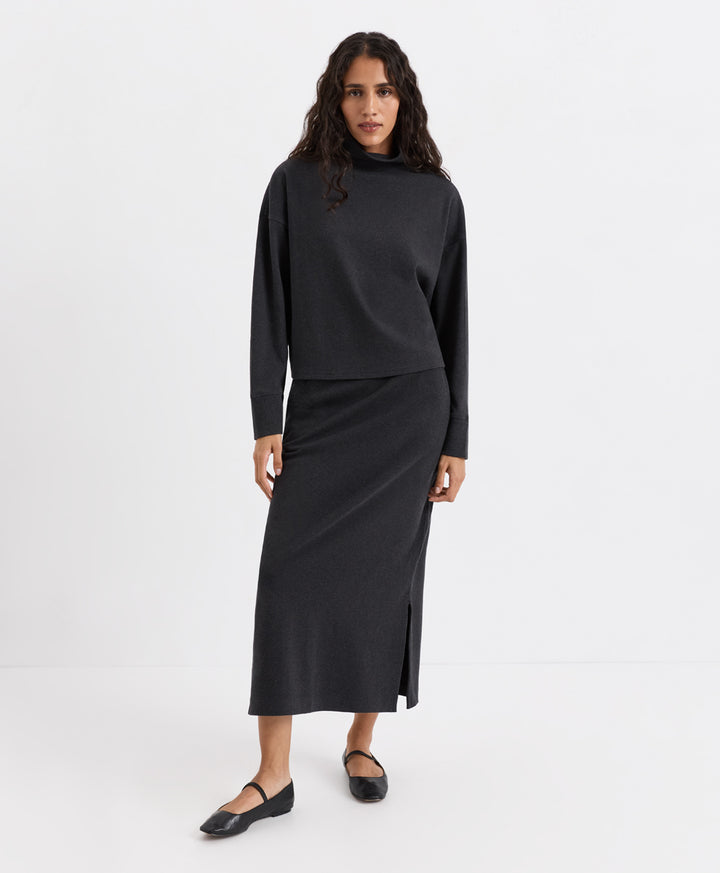 A woman modeling a dark charcoal heather two-piece knit set consisting of a long-sleeve funnel-neck top and a matching midi skirt with a front slit, paired with black flats, against a plain white studio background.