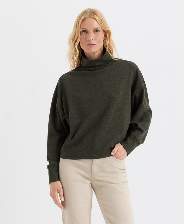 A woman modeling a loden green funnel-neck top with long sleeves and a relaxed fit, paired with beige pants, against a plain light gray studio background.