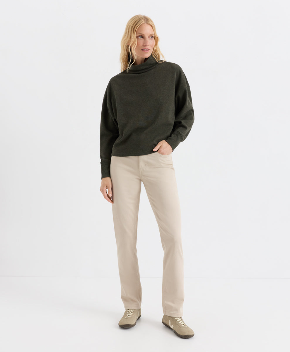 A woman modeling a loden green funnel-neck top and beige straight-leg trousers, paired with beige shoes, against a clean white studio background.