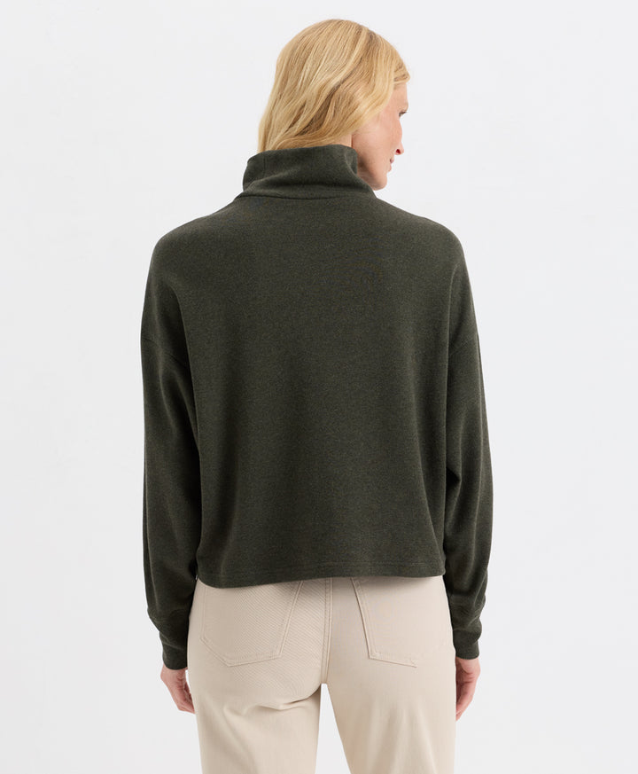 A woman shown from the back wearing a loden green funnel-neck top and light beige trousers, against a plain white studio background.