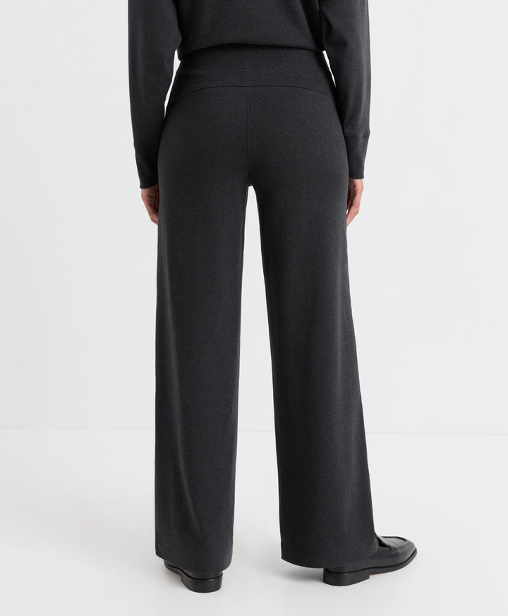 A person shown from the back wearing dark charcoal wide-leg trousers, a matching dark long-sleeve top, and black shoes against a clean white studio background.