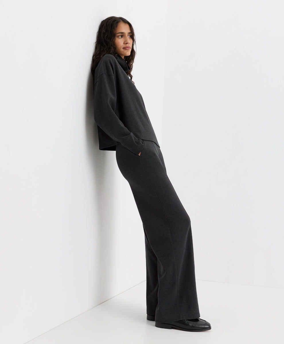 A woman wearing a dark charcoal heather long-sleeve top and matching wide-leg trousers, leaning against a white wall in a minimalist studio setting.