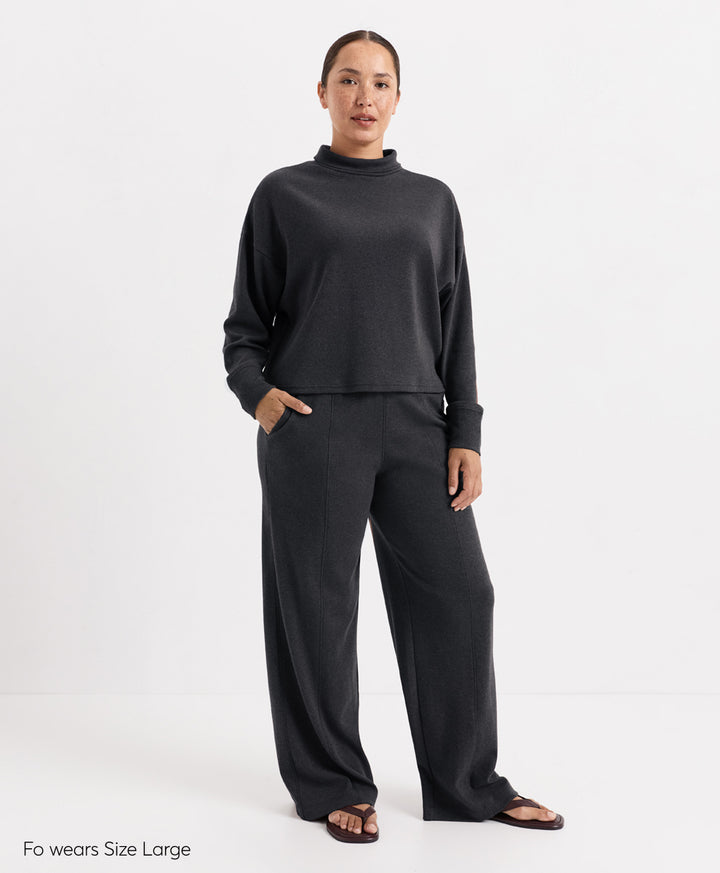 A woman modeling a dark charcoal heather knit two-piece set consisting of a cropped mock-neck long-sleeve top and matching wide-leg pants, standing against a plain white studio background.