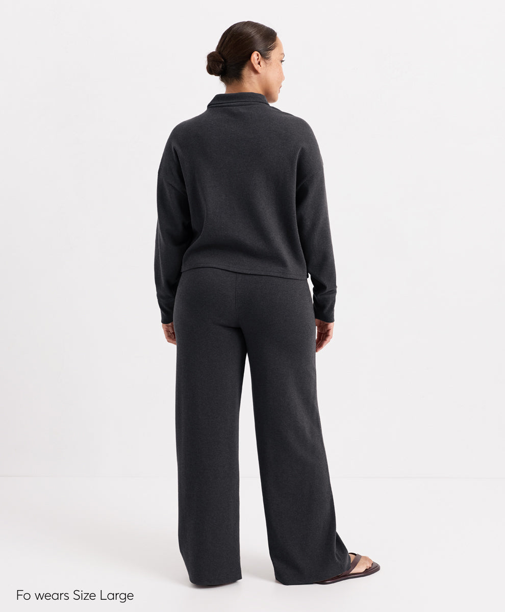 A model shown from the back wearing a dark charcoal heather mock-neck long-sleeve top and matching wide-leg trousers, photographed against a clean white studio background.