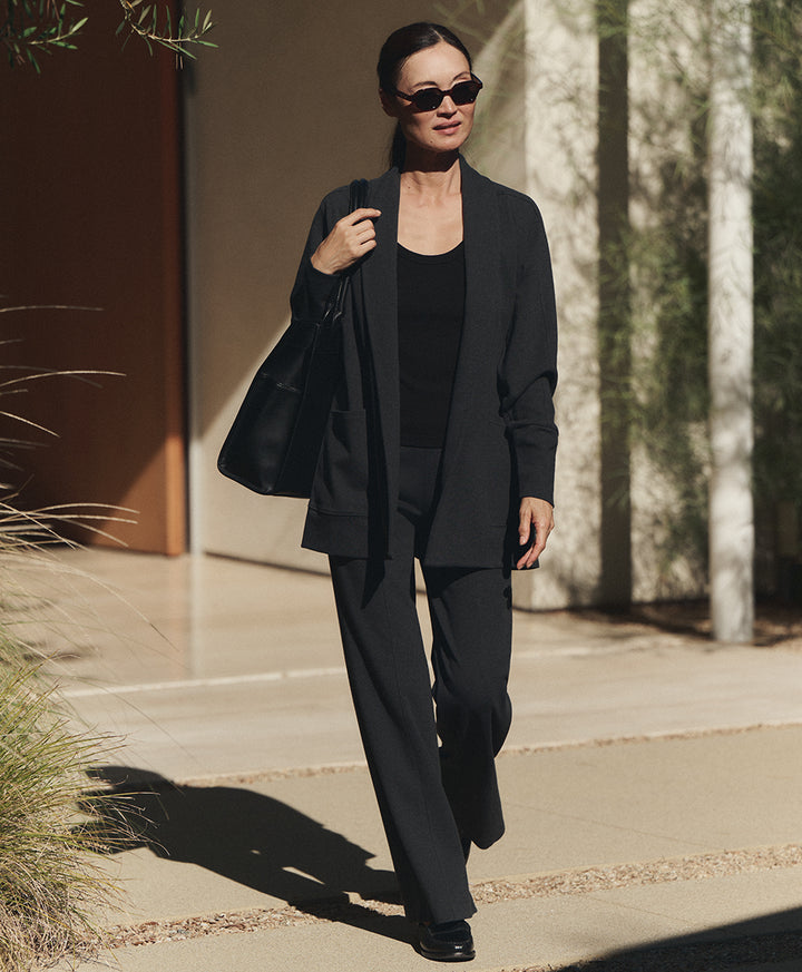 A woman walking outdoors wearing a cardigan, dark charcoal heather wide-leg trousers, a black top, black sunglasses, and carrying a black shoulder bag.