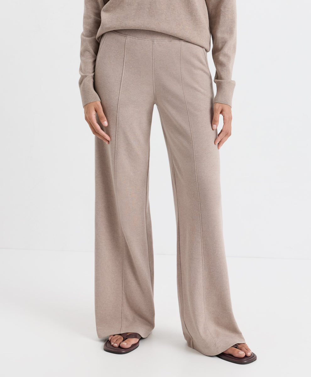 A person wearing high-waisted, wide-leg knit pants in a light taupe-beige color, paired with a matching long-sleeve top and dark brown slide sandals, photographed against a clean white studio background.