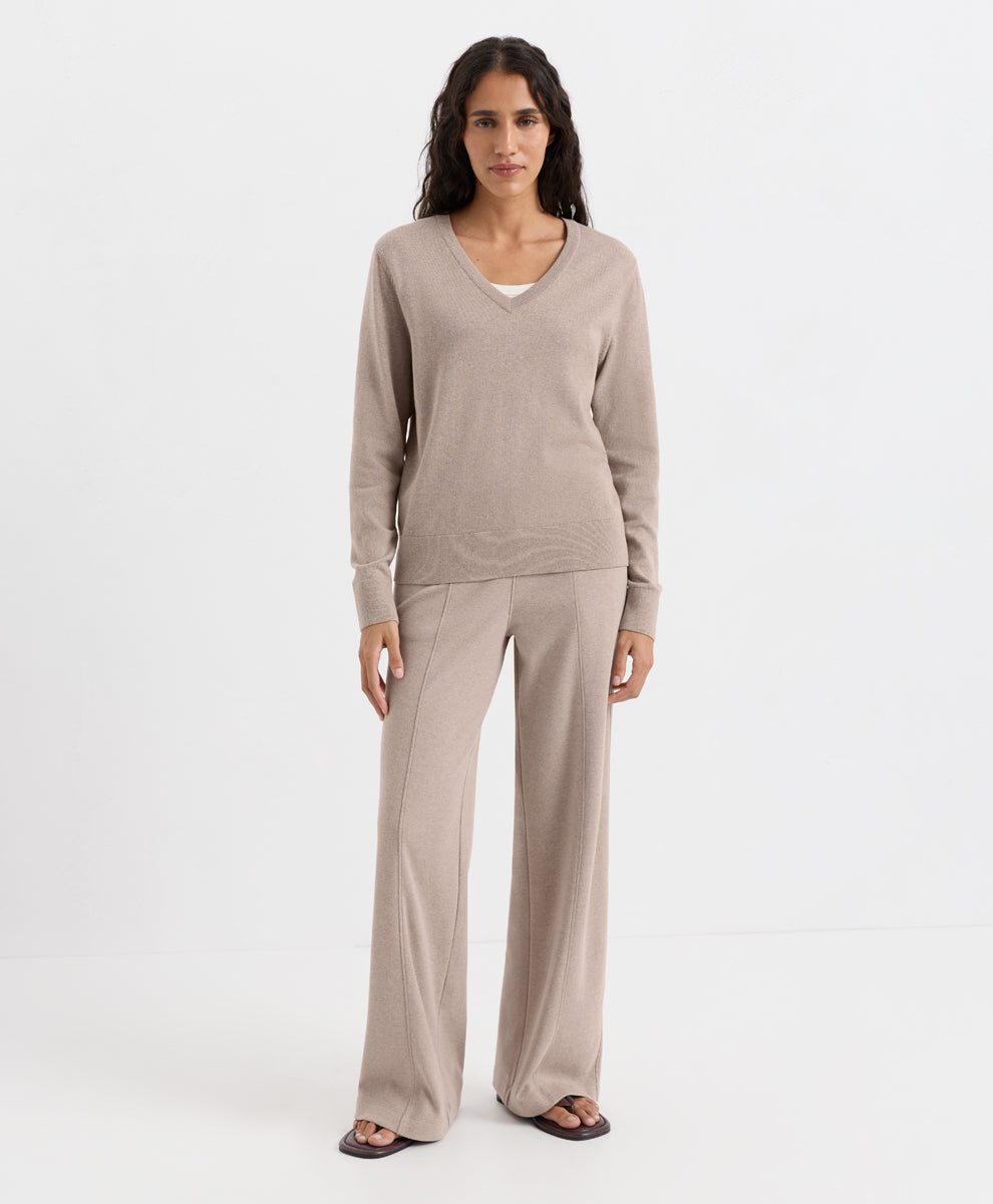 A woman modeling a matching lounge set consisting of a long-sleeve V-neck knit top and wide-leg pants in a light taupe-beige color, paired with dark sandals, against a plain white studio background.