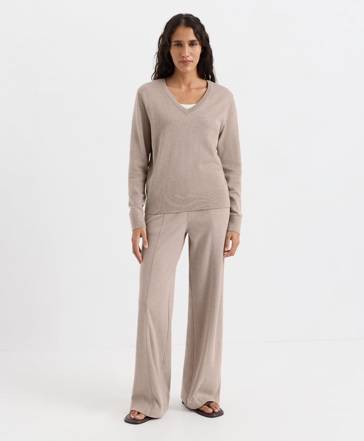 A woman modeling a matching lounge set consisting of a long-sleeve V-neck knit top and wide-leg pants in a light taupe-beige color, paired with dark sandals, against a plain white studio background.