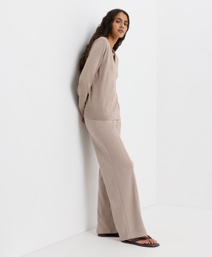 A woman wearing a matching beige knit lounge set consisting of a long-sleeve top and wide-leg pants, leaning against a white wall in a minimalist studio setting.