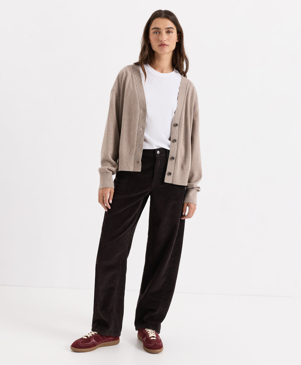 A woman stands against a clean white studio background wearing a light taupe cardigan over a white top, espresso corduroy barrel pants, and burgundy suede shoes.