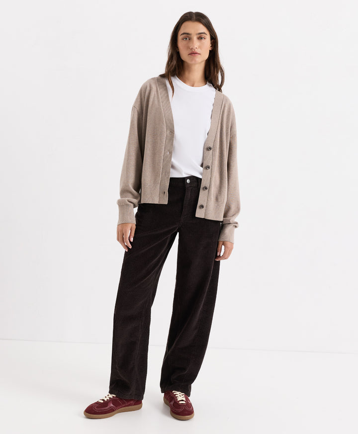 A woman stands against a clean white studio background wearing a light taupe cardigan over a white top, espresso corduroy barrel pants, and burgundy suede shoes.