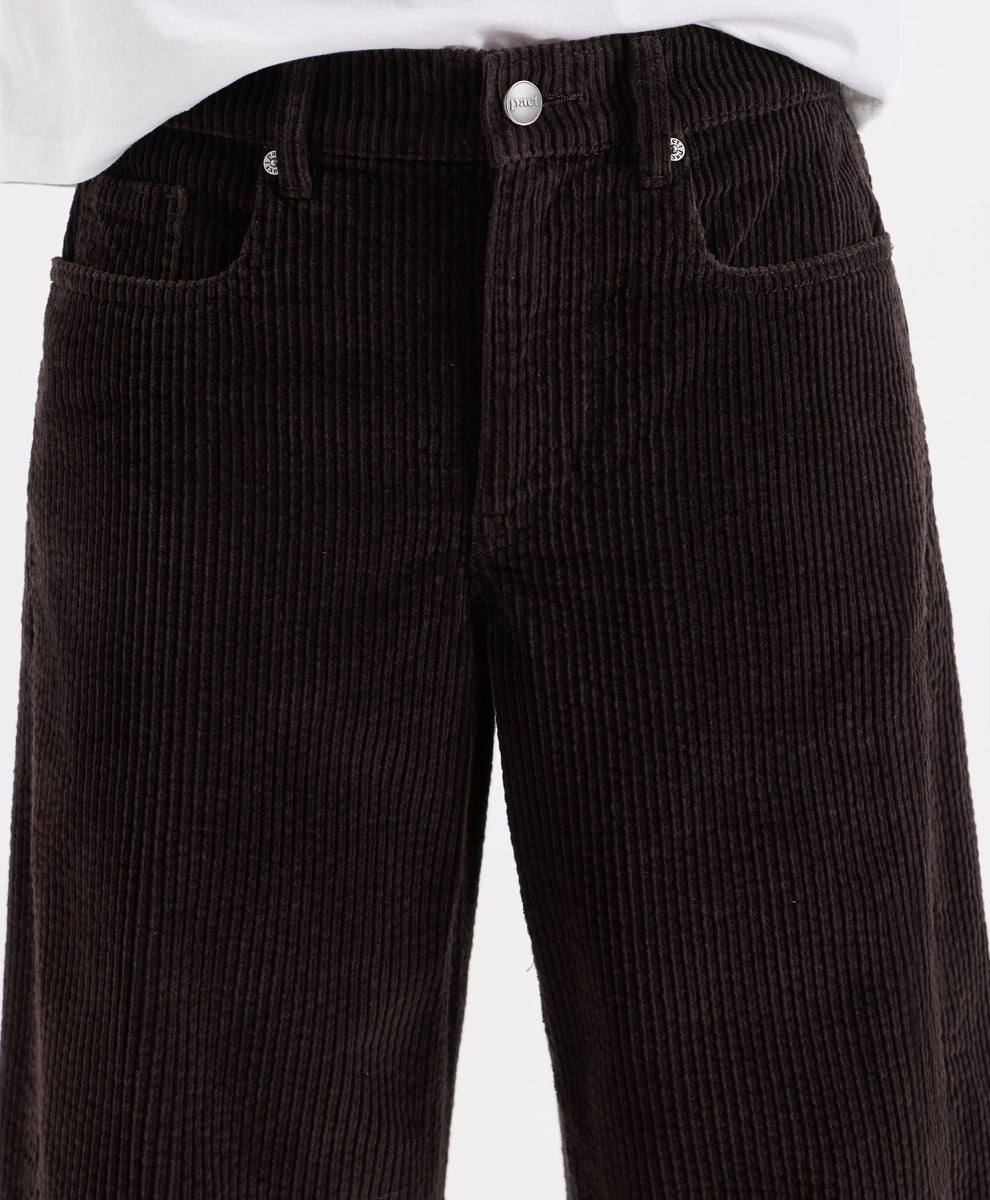 Close-up of espresso corduroy barrel pants with a white shirt hem visible at the top, showing the front waistband, button, belt loops, and side pockets.