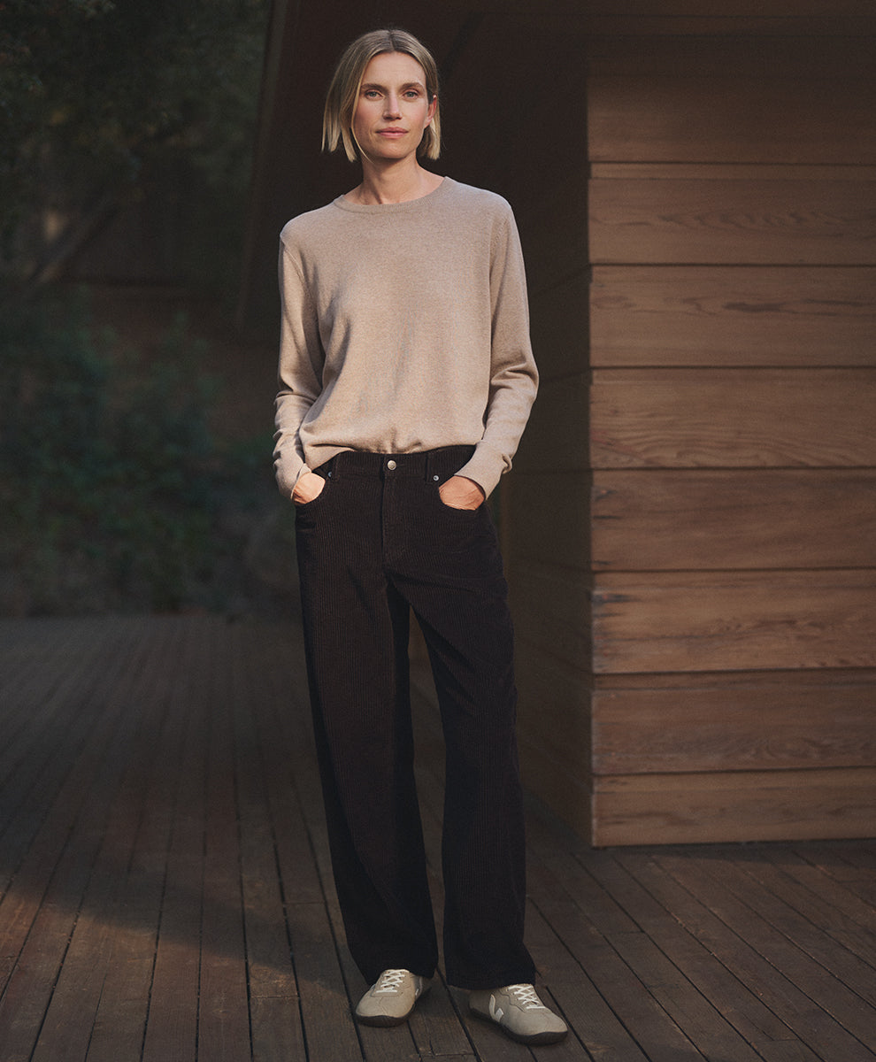 A woman stands on a wooden deck wearing a light beige crewneck sweater, espresso barrel pants, and white sneakers, with a wooden wall and dark background behind her.