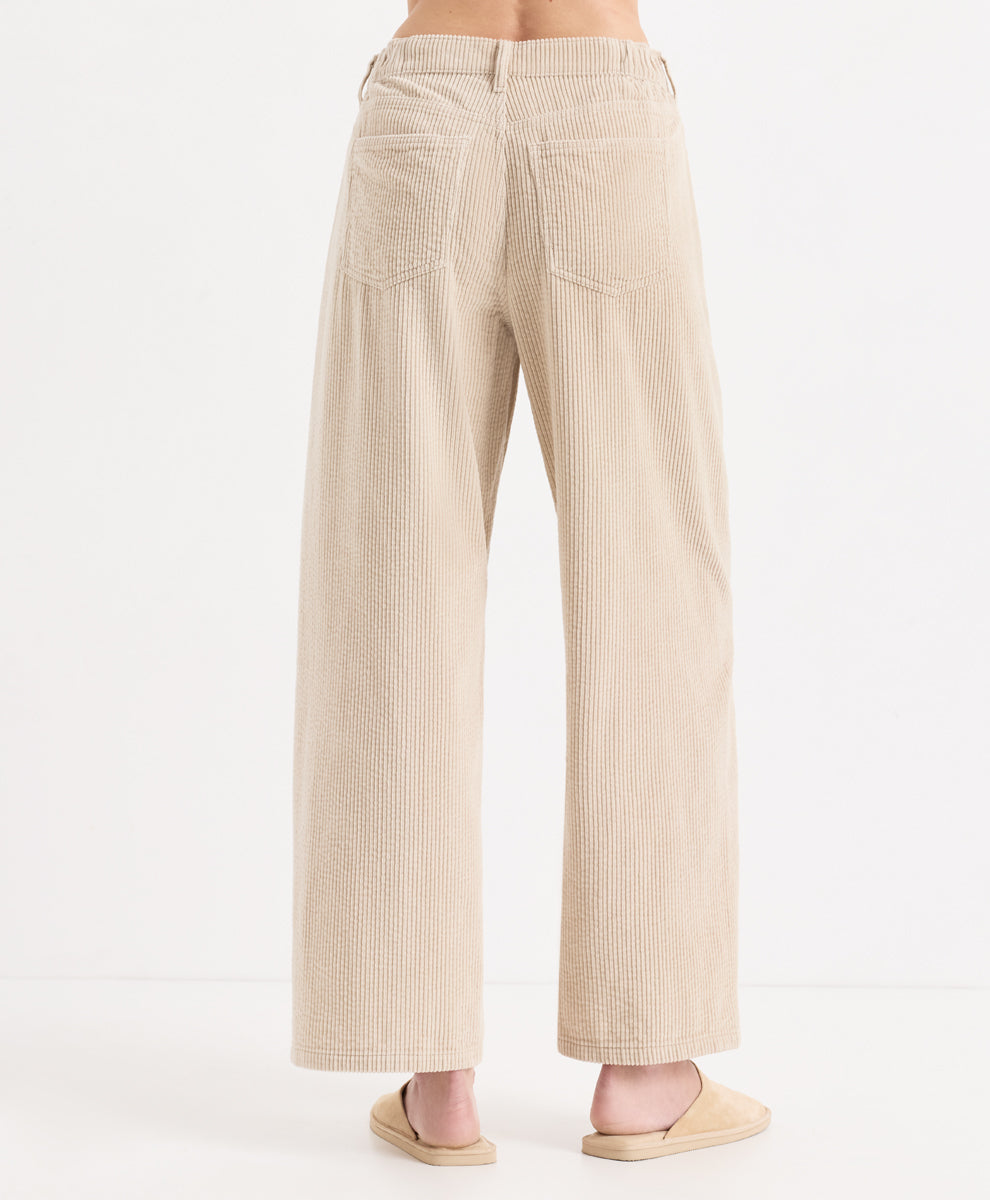 Back view of a person wearing high-waisted, corduroy barrel pants in parchment, paired with matching beige slide sandals, against a plain white studio background.