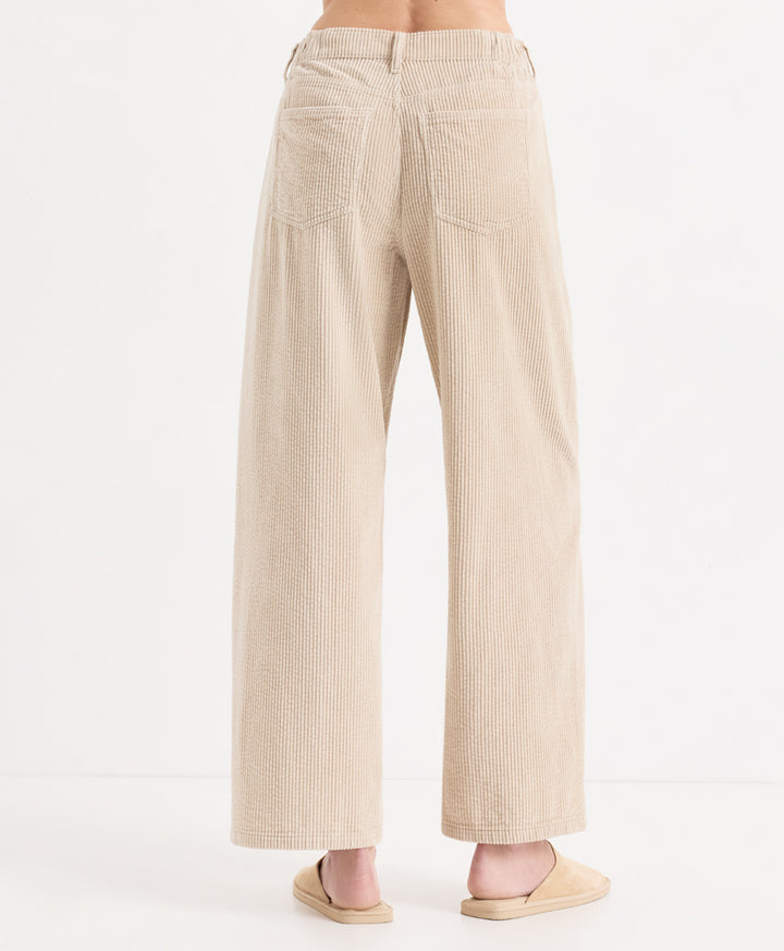 Back view of a person wearing high-waisted, corduroy barrel pants in parchment, paired with matching beige slide sandals, against a plain white studio background.