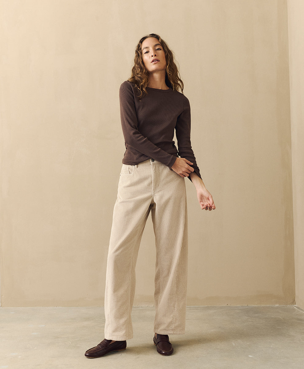 A woman modeling a dark brown long-sleeve boat-neck knit top and parchment barrel pants, standing against a neutral beige plaster wall.