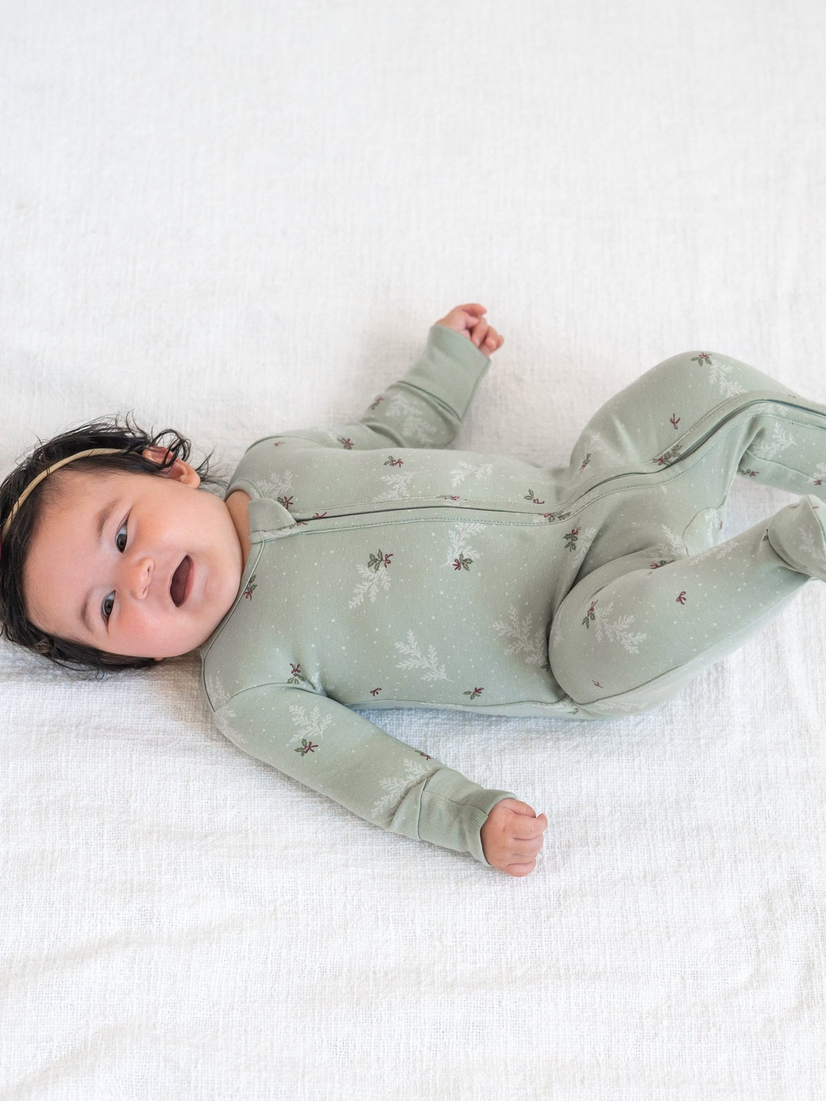 A baby lying on a white textured blanket, wearing a sage green footed sleeper with a subtle brown and white pattern.