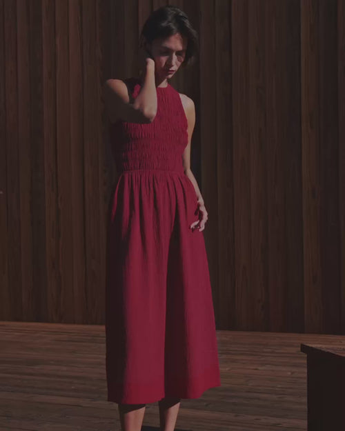 Woman wearing Pact's Organic Coastal Double Gauze Smocked Dress in Poppy Red standing against a wood panel wall.