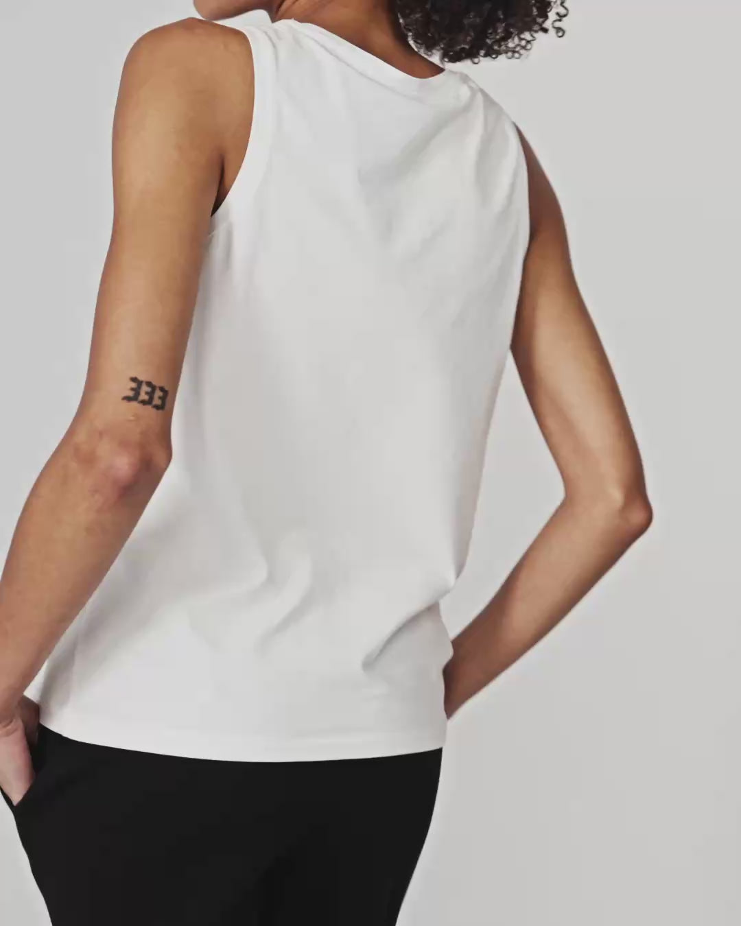 Woman wearing a white Pact Organic Luxe Essential Scoop Neck Tank and dark pants against a white background