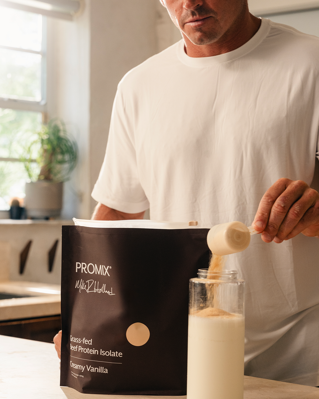 A man in a white t-shirt is shown in a kitchen setting, pouring protein powder from a scoop into a glass bottle. In the foreground, a brown bag of PROMIX Whey Isolate in Creamy Vanilla flavor is clearly visible.