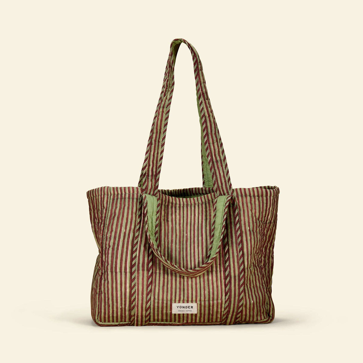 A striped cotton tote bag with vertical wavy lines in brown, beige, and muted green, featuring light green fabric handles and a small white brand label at the bottom front.
