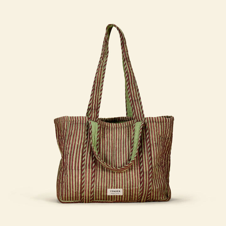 A striped cotton tote bag with vertical wavy lines in brown, beige, and muted green, featuring light green fabric handles and a small white brand label at the bottom front.
