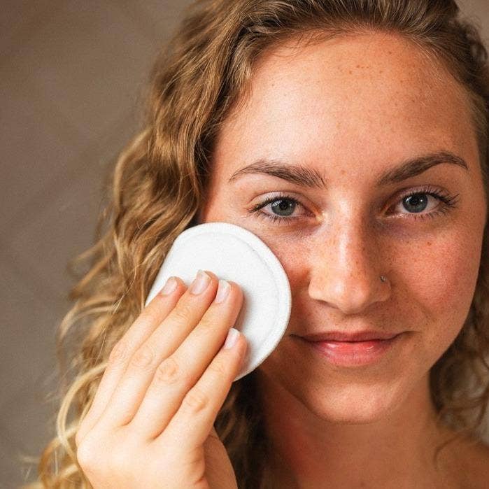 Woman using a white bamboo round on her face against a neutral background