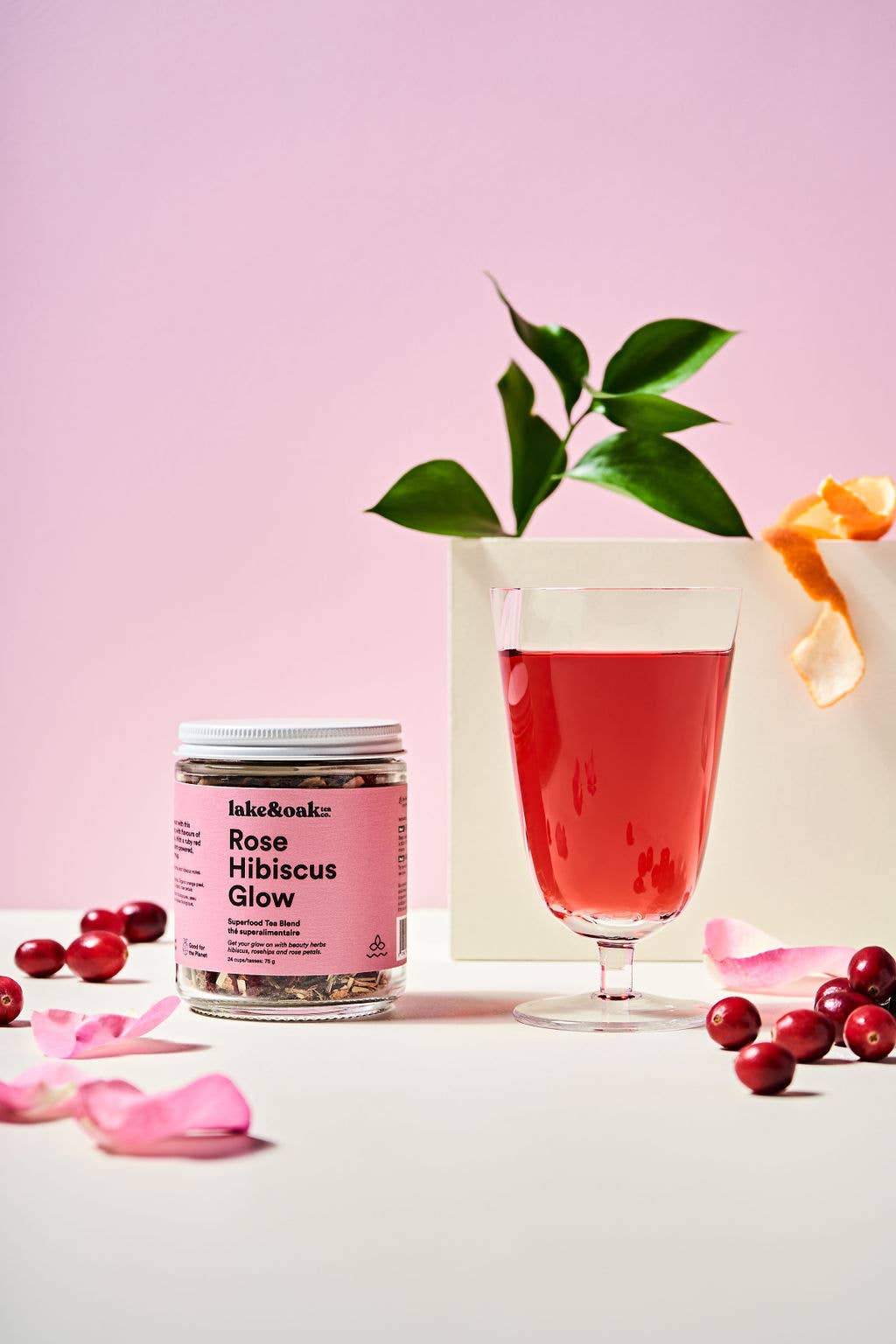 Jar of 'Rose Hibiscus Glow' product with a glass of pink drink and cranberries on a light pink background