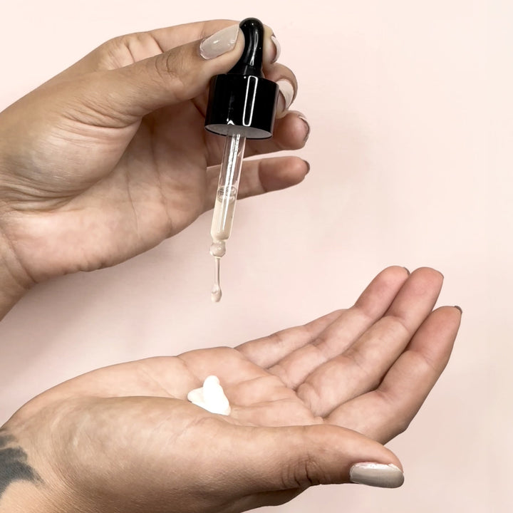 Hand holding a dropper with a clear liquid over another hand  with lotion on a pink background
