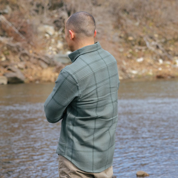 A man standing by a river, viewed from behind, wearing a lichen plaid long-sleeve shirt and tan pants.